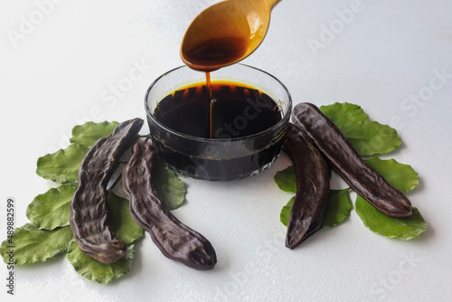 Photos carob molasses in glass bowl and in wooden spoon and carob pods on rustic backgr