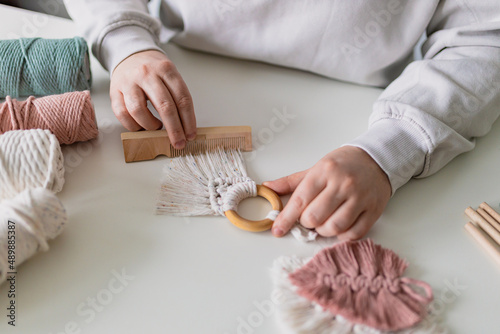Obraz na płótnie Woman working out last details of an almost-finished macrame piece, combing ropes inside a home room. Close up. Natural cotton threads and wooden beads. Female hobby. Wall hanging decor. w salonie