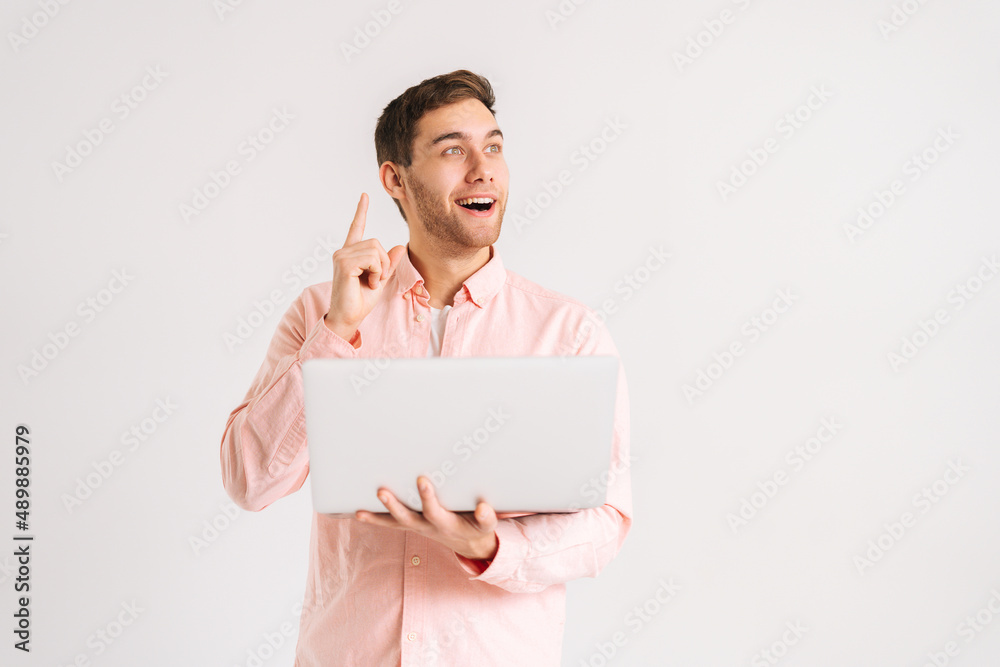 Portrait of amazed young man standing with laptop computer pointing index finger up with happy face, looking away on white isolated background. Studio shot of happy male student posing with notebook. Portrait of amazed young man standing with laptop computer pointing index finger up with happy face, looking away on white isolated background. Studio shot of happy male student posing with notebook.