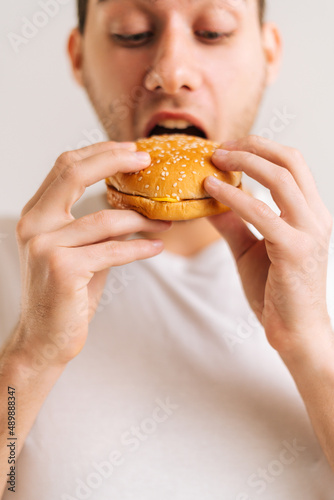 Vertical cropped shot of handsome young man with enjoying eating delicious burger on white isolated background. Close-up studio portrait of happy handsome male holding tasty unhealthy hamburger.