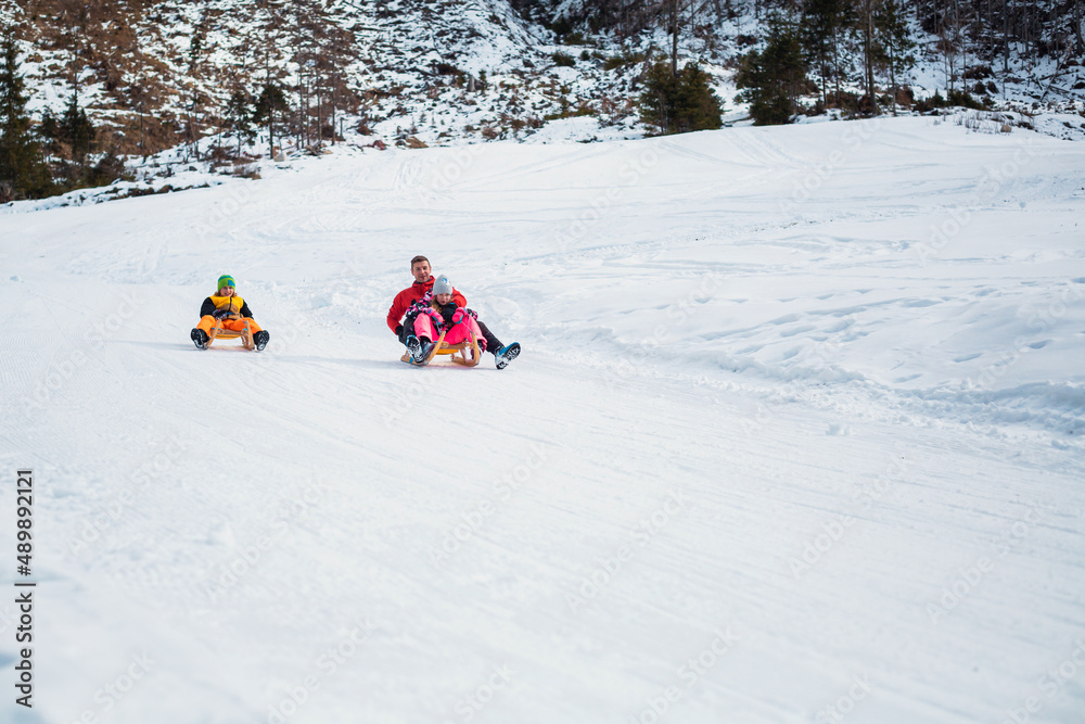 Cheerful Caucasian boy and girl enjoying with father in winter play, sliding down sleds on a snow covered slope.