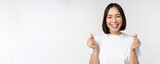 Beautiful asian woman smiling, showing finger hearts gesture, wearing tshirt, standing against white background