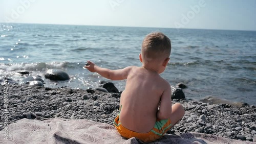 A boy-child sits with his back to the camera and throws a small pebble into the sea. Vacation on the seashore with the whole family.