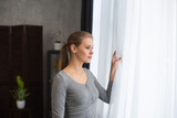 Young teenage girl is standing in front of the window and looking into it. Blond woman at home.
