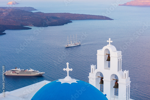 White church with blue dome roof overlooking cruise ships on Aegean sea, Santorini, Greece.