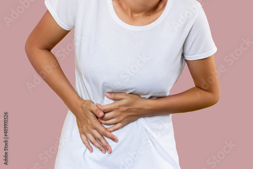 Cropped shot of a young woman in a white t-shirt holding her stomach with her hands isolated on pastel color background. Stomach pain