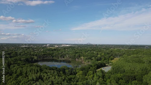 Aerial view over Lake Minnetonka with clear blue sky with wispy clouds. Lush green foliage all around. Water from the lake reflecting the sun.  Park tucked into forest. 