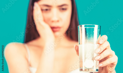 Woman taking drugs to releave headache. Brunette take some pills, holds glass of water, isolated on blue. Young woman taking pill against headache. Brunette taking a pill with a glass of water photo