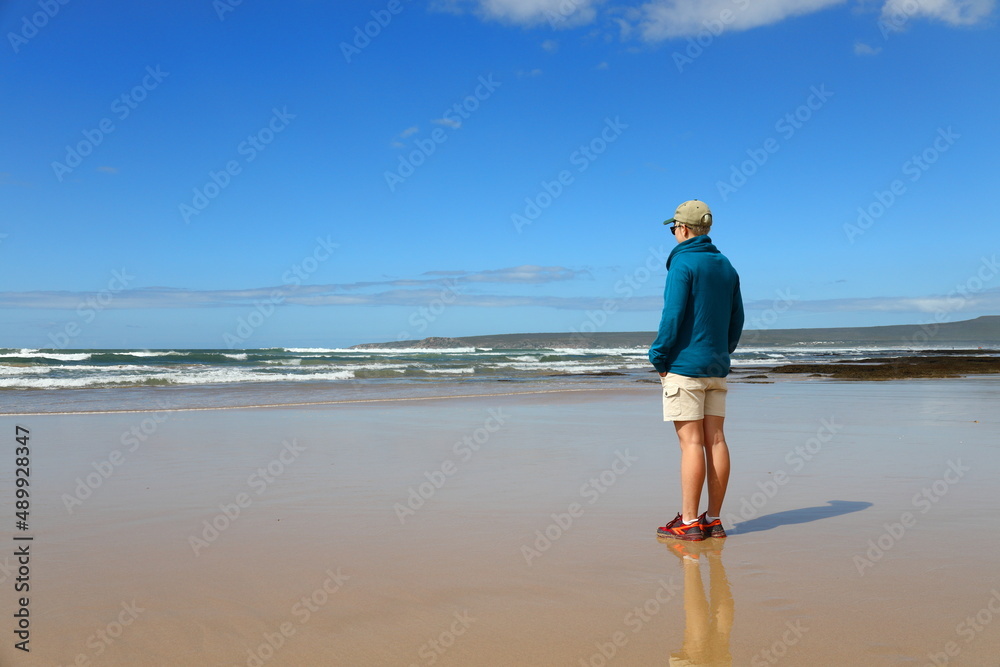 A young, blonde woman standing on the beach and looking out to sea.