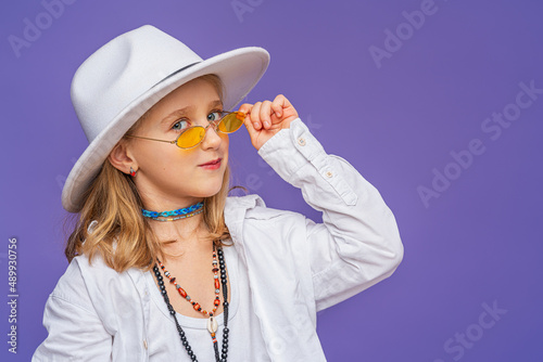 Portrait of a happy cute little girl in a hat and unisex clothes on a purple background in full growth. A stylish children's model is having fun and feels stunned, amazed.