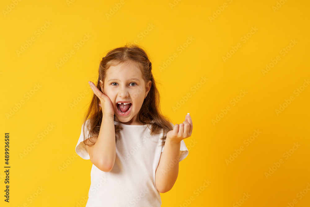 cute smiling preschool girl holds her first fallen baby tooth in her hand while standing on yellow background. growing permanent tooth is in the open mouth. . The concept of hygiene of baby teeth. cute smiling preschool girl holds her first fallen baby tooth in her hand while standing on yellow background. growing permanent tooth is in the open mouth. . The concept of hygiene of baby teeth.