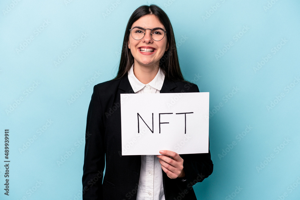 Young caucasian woman holding a NFT placard isolated on blue background laughing and having fun. Young caucasian woman holding a NFT placard isolated on blue background laughing and having fun.