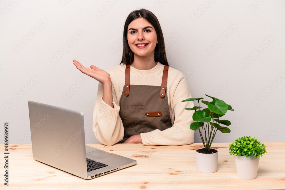 Young caucasian gardener woman working on the table isolated on pink background showing a copy space on a palm and holding another hand on waist. Young caucasian gardener woman working on the table isolated on pink background showing a copy space on a palm and holding another hand on waist.
