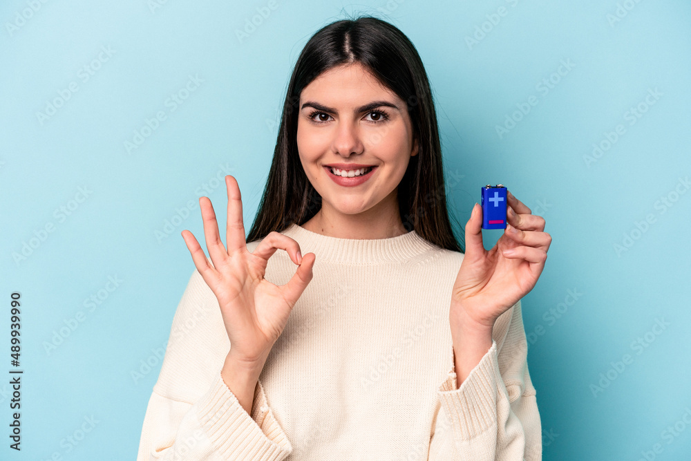 Young caucasian woman holding a batterie isolated on blue background cheerful and confident showing ok gesture. Young caucasian woman holding a batterie isolated on blue background cheerful and confident showing ok gesture.