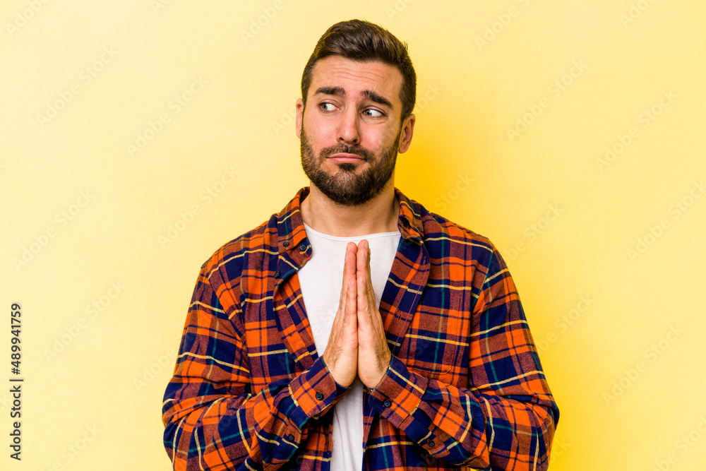 Young caucasian man isolated on yellow background praying, showing devotion, religious person looking for divine inspiration.