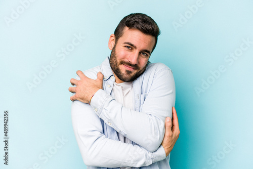 Young caucasian man isolated on blue background hugs, smiling carefree and happy.