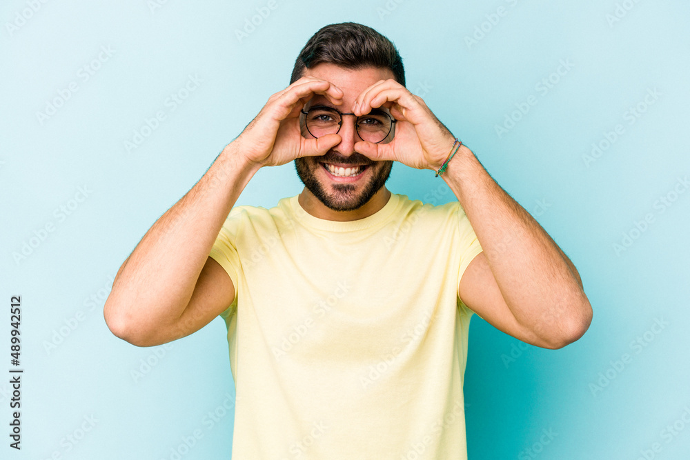 Young caucasian man isolated on blue background showing okay sign over eyes Young caucasian man isolated on blue background showing okay sign over eyes