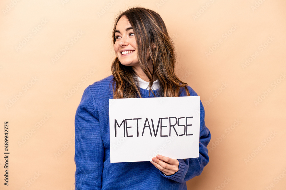 Young caucasian woman holding a metaverse placard isolated on beige background looks aside smiling, cheerful and pleasant.
