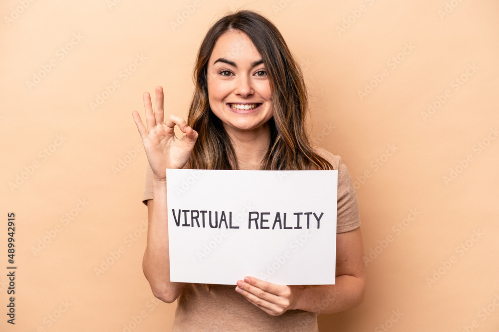 Young caucasian woman holding a virtual reality placard isolated on beige background cheerful and confident showing ok gesture. Young caucasian woman holding a virtual reality placard isolated on beige background cheerful and confident showing ok gesture.
