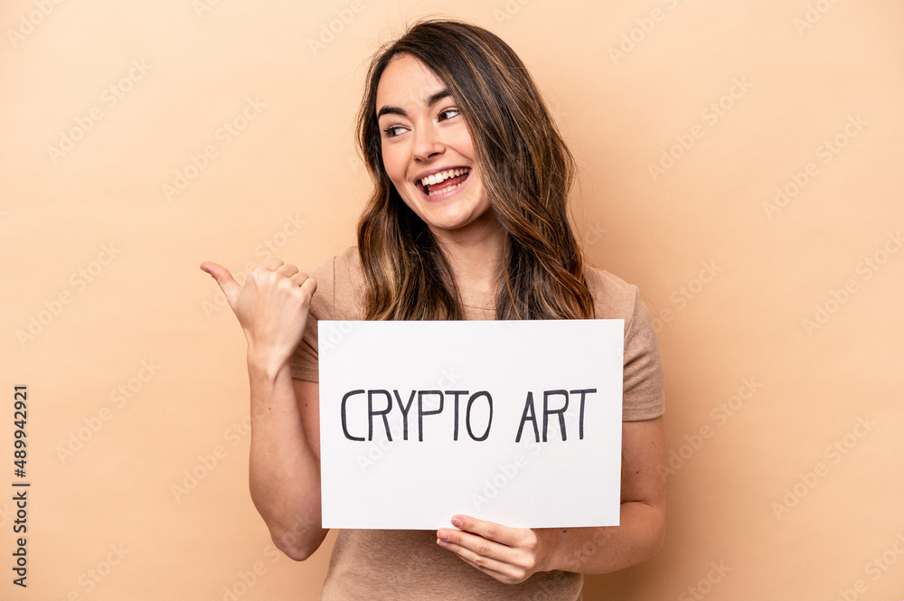Young caucasian woman holding a crypto art placard isolated on beige background points with thumb finger away, laughing and carefree. Young caucasian woman holding a crypto art placard isolated on beige background points with thumb finger away, laughing and carefree.