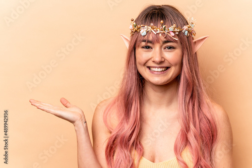 Young elf woman with pink hair isolated on beige background showing a copy space on a palm and holding another hand on waist.