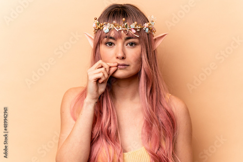 Young elf woman with pink hair isolated on beige background with fingers on lips keeping a secret. © Asier