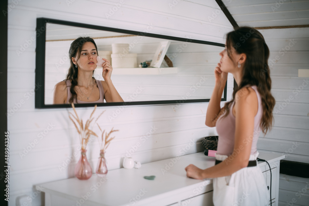 A young woman makes a facial massage with a gouache scraper at the mirror at home. A young woman makes a facial massage with a gouache scraper at the mirror at home.