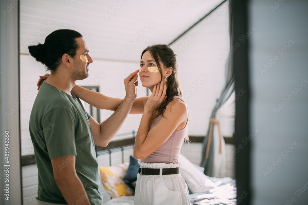 Naklejka premium Family spa day at home. Young woman and man with golden hyaluronic patches on their faces in the bedroom of their home.