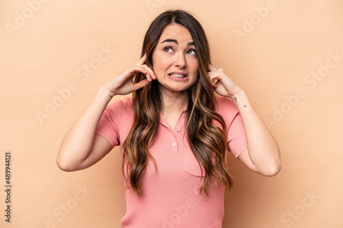 Young caucasian woman isolated on beige background covering ears with fingers, stressed and desperate by a loudly ambient.