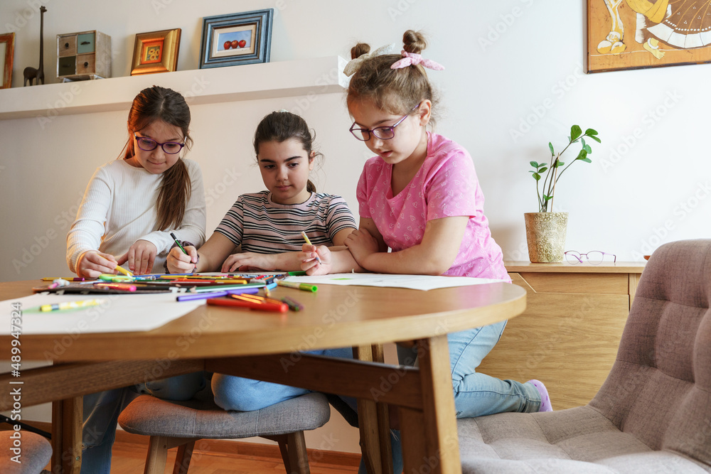 A group of school girls draw with color pencils. They're sitting at the table in the living room and having fun. A group of school girls draw with color pencils. They're sitting at the table in the living room and having fun.