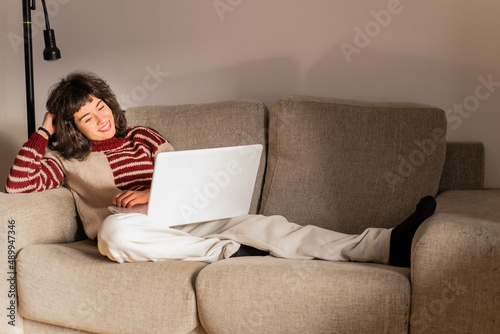 A mid adult brunette Spanish woman is watching a film on her laptop lying on her sofa at home, wearing comfy clothes, indoors, red and grey 