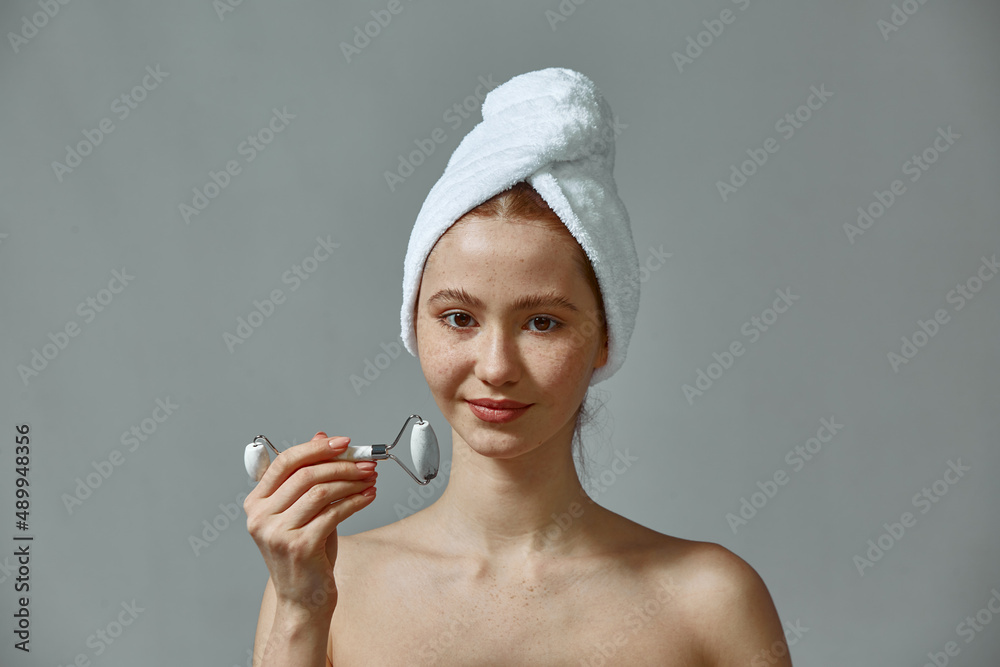 Young girl hold massager jade roller for facial massaging. Female with bare shoulders, towel on head show face massage Young girl hold massager jade roller for facial massaging. Female with bare shoulders, towel on head show face massage