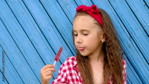 A girl in a red plaid shirt and a red bow in her hair licks a sweet lollipop. Excessive consumption of sweets and sugar is harmful to health. 