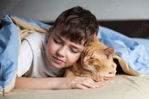 The boy hugs a red cat on a bed under a blanket