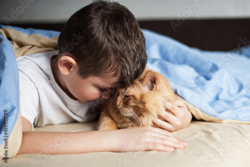The boy hugs a red cat on a bed under a blanket