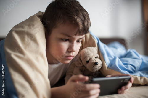 a boy with a toy lies under a blanket and plays on the phone