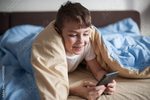 boy of European appearance lies under a blanket with a phone in his hands and communicates with a smile in an online chat
