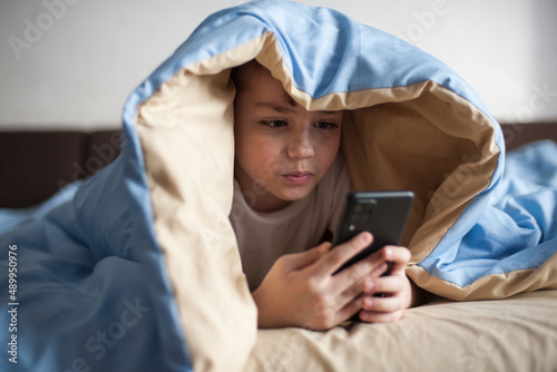 The boy lies under the covers with the phone