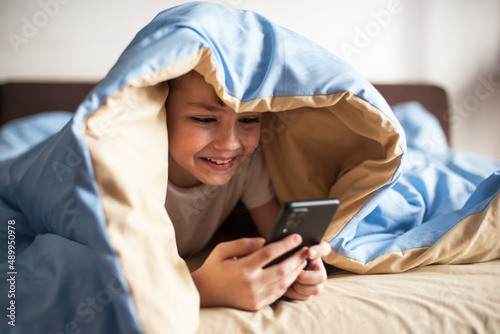 boy of European appearance lies under a blanket with a phone in his hands and communicates with a smile in an online chat