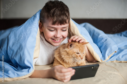 a boy and a red cat look at the phone lying under a blanket