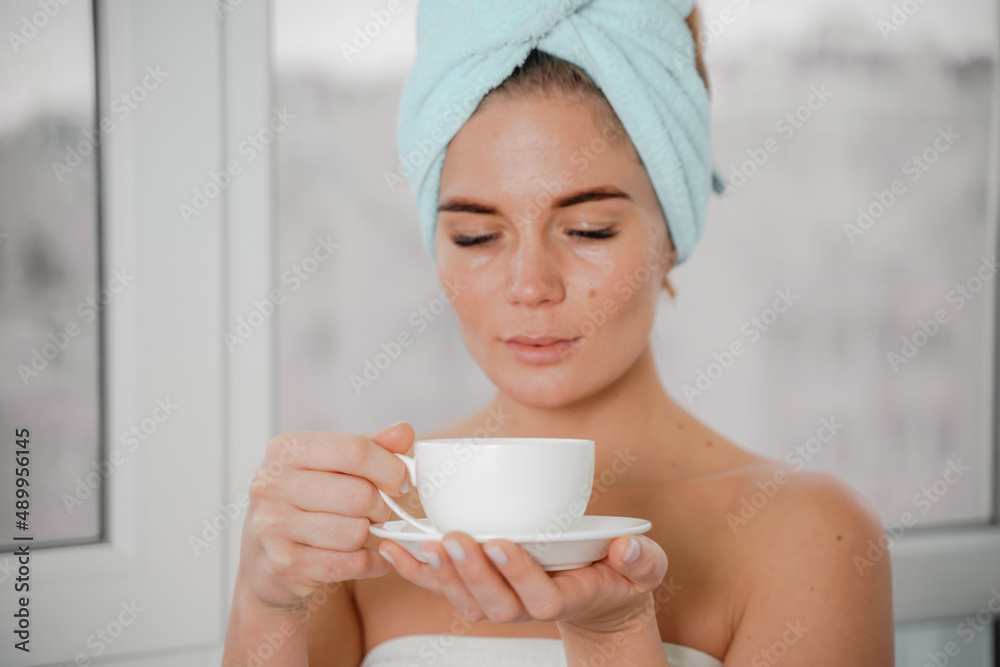 Young serene relaxed woman in spa bath towel drinking hot beverage tea coffee after taking shower bath at home. Beauty treatment, hydration concept.