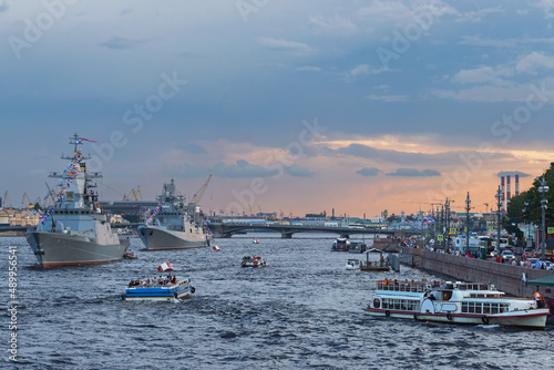 small boats with tourists inspect warships on the Neva in the city center against the blue evening sky