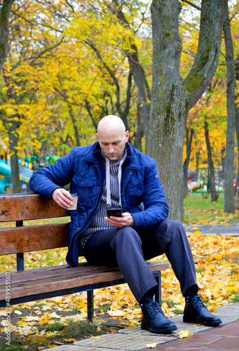a young man sits on a bench in an autumn park and looks into a smartphone, holding a cup of coffee or tea in his other hand. wireless technologies development concept