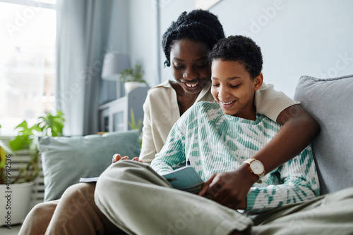 Portrait of happy black mother with daughter doing homework together while sitting on sofa at home