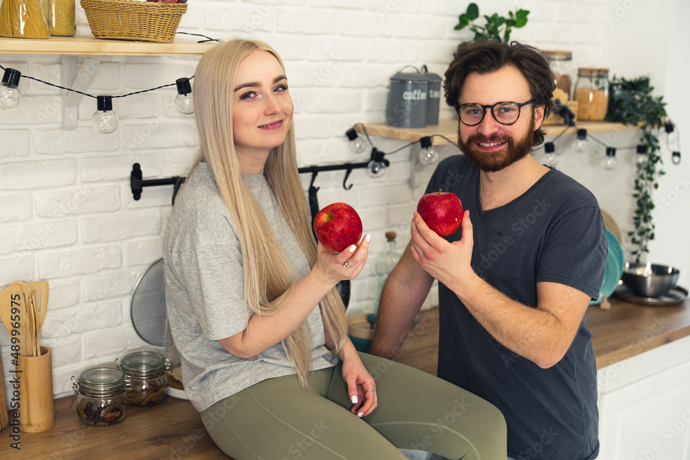 Obraz premium young married caucasian couple sharing healthy lifestyle, holding a red apple while being in the kitchen and looking at camera. High quality photo