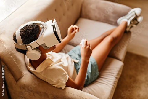 High angle portrait of African American girl playing VR game while laying on sofa at home photo