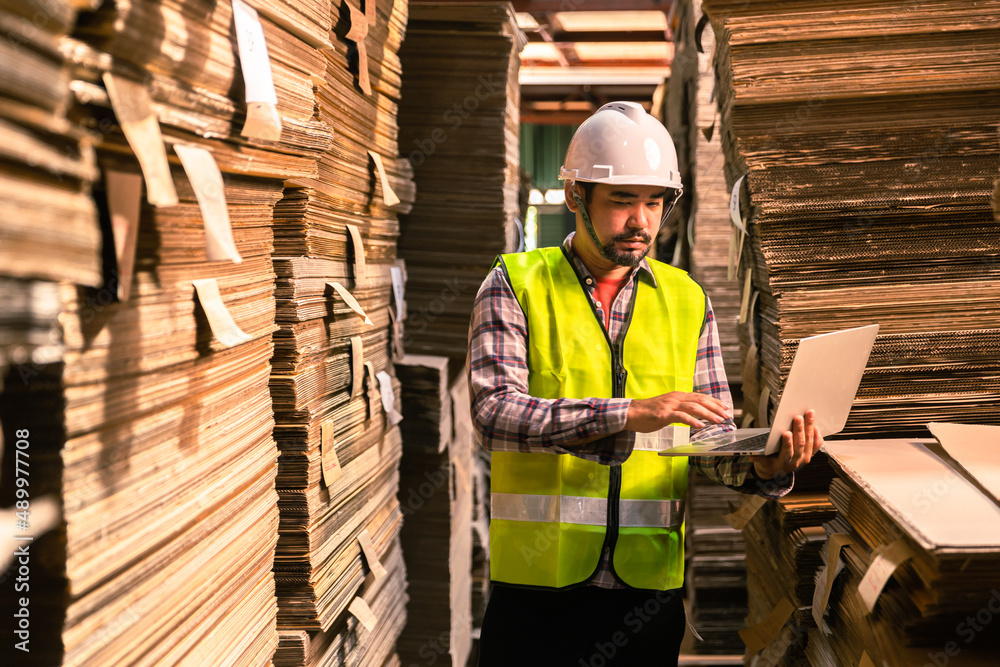 Engineer man wearing safety helmet and vest holding clipboard and take note on the paper in the automotive part warehouse.Products and corrugated cardboard. 