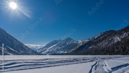 The frozen lake is surrounded by picturesque mountains. Tire tracks are visible on the snow-covered surface.The sun is shining in the blue sky. Altai. Lower Multinskoe Lake.