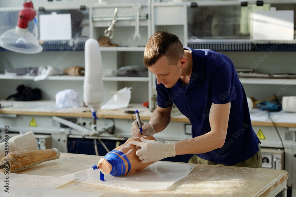 Worker of manufactory with blue highlighter bending over workbench while putting marks on silicone liner of leg prosthesis Worker of manufactory with blue highlighter bending over workbench while putting marks on silicone liner of leg prosthesis