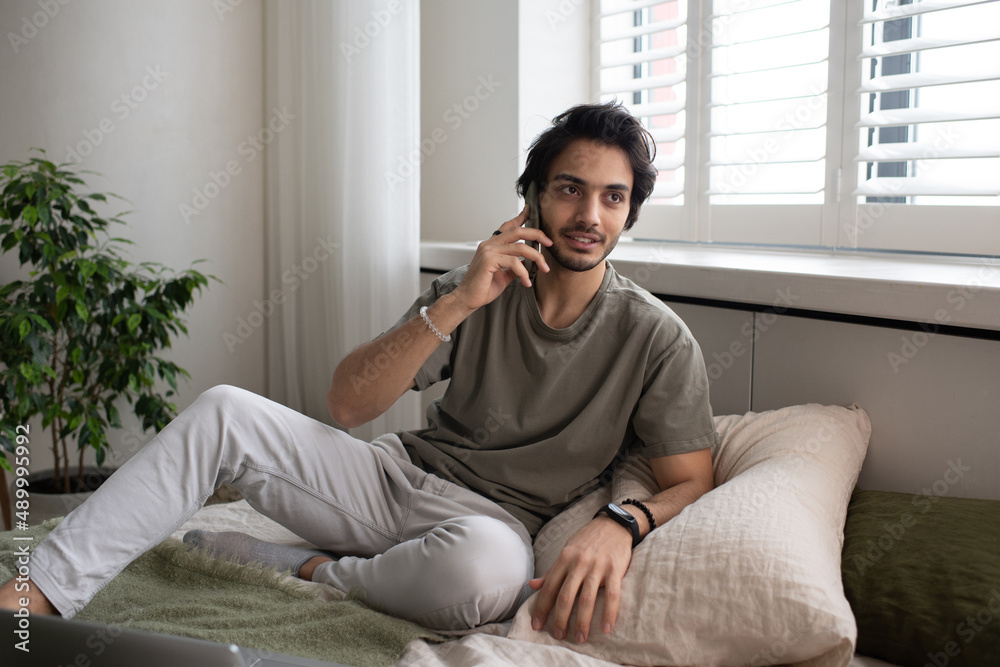 Young contemporary male employee or freelancer talking on mobile phone while sitting on bed against window